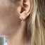 a model with hair tucked behind her ear is shown wearing a pair of gold ball stud earrings with dangling teardrop faceted aquamarine gemstones