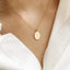 Model is wearing a delicate gold chain necklace with a small, oval gold Mother Mary pendant showing the back of the charm featuring an M with a cross above surrounded by tiny stars and dainty hearts.