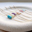 Six dainty birthstone bracelets lined up on a stoneware dish showing three turquoise roundels surrounded on each end by two gold beads lined up on a delicate cable chain. The bracelets are turquoise, opal, amethyst, pink opal, pink tourmaline and aquamarine.
