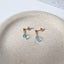 a pair of gold ball stud earrings with dangling teardrop faceted aquamarine gemstones displayed on a round stoneware dish