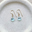 A pair of blue topaz stones dangling from a gold ear wire displayed on a stoneware jewelry dish