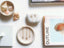 A collection of summer jewelry that includes dainty summer inspired charms and earrings including a sun charm necklace, starfish charm necklace, anchor charm necklace, and mother of pearl starfish charm threader earrings all displayed on a jewelry dish with a collection of shells and a book relating to summer 