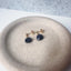 Dainty blue sapphire stud earrings displayed in a beige ceramic jewelry dish.
