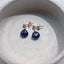 Dainty blue sapphire teardrop stud earrings with gold settings displayed in a neutral ceramic dish.