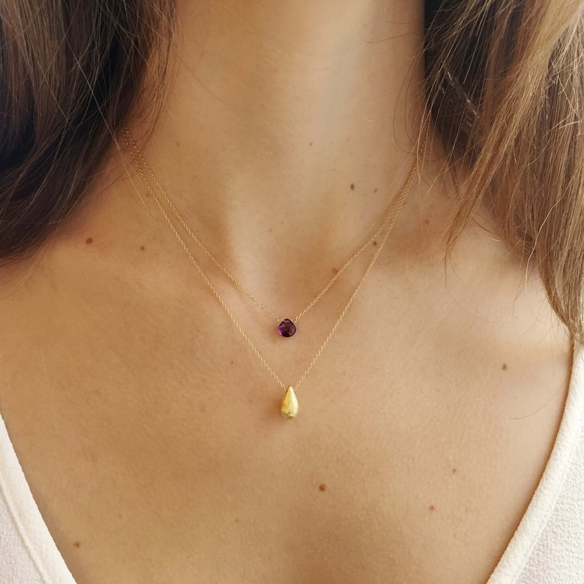 A model is shown wearing a layered necklace with a genuine purple amethyst layered with a gold teardrop pendant