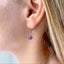 model is shown wearing an amethyst birthstone earring that dangles from a simple gold ear wire