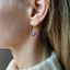 model is shown wearing an amethyst birthstone earring that dangles from a simple gold ear wire