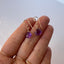 model is shown holding in her hand a pair of amethyst birthstone earrings attached to simple gold ear wires with a tiny coil
