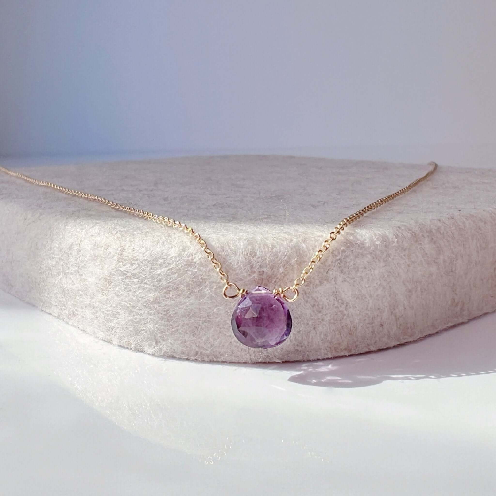 A dainty amethyst birthstone necklace on a fine gold cable chain, displayed on a beige cushion.