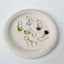 a collection of dainty birthstone drop earrings displayed on a stoneware jewelry dish showing peridot, blue topaz, amethyst, garnet, citrine and crystal birthstones.