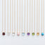 Nine delicate gold chain necklaces with small teardrop-shaped gemstone pendants are lined up horizontally against a clean white background. The pendants are in different colors: burgundy, clear, pale pink, light blue, purple, green, yellow, orange, and turquoise.